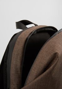 Sac à dos en tissu marron avec un motif en chevrons, comportant des accents et des fermetures éclair noirs, montrant l'intérieur et une ouverture partielle.