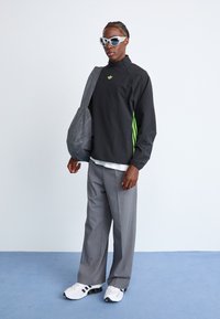 Black windbreaker with green accents, gray bag, gray trousers, and white sneakers. Accessorized with oversized white sunglasses.