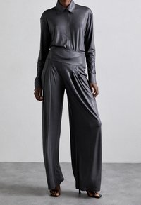 Person klädd i en mörkgrå långärmad skjorta med krage instoppad i högmidjade vida satinsbyxor med svarta fyrkantiga klackar.