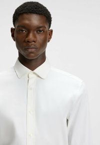 White long-sleeve shirt made of smooth fabric with a pointed collar and a buttoned front placket. Features minimalistic design and soft texture.