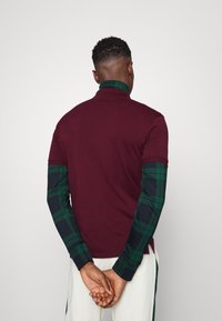 Burgundská polo košile s dlouhými rukávy v zeleno-černém kostkovaném vzoru. Směs bavlny, uvolněný střih, detaily límce a viditelné švy na lemování.