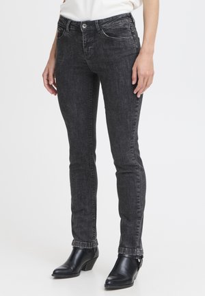 Vaqueros gris oscuro de corte ajustado con botines negros y una camiseta blanca, mostrados desde la cintura hasta los pies sobre un fondo neutro.