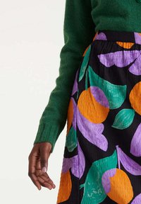 Mano de piel oscura al lado de una falda con grandes patrones florales en naranja, morado y verde, combinada con un suéter verde de manga larga.