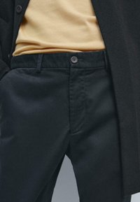 Pantalon noir avec une texture lisse, dotée d'une fermeture à bouton, de poches latérales et d'une coupe ajustée, associé à un haut jaune clair.