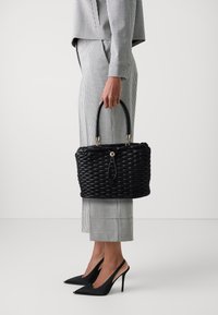 Borsa a tracolla nera in tessuto intrecciato, con una forma strutturata, dotata di dettagli in metallo dorato e di un manico rotondo. Abbinata a un outfit a quadri e tacchi neri.