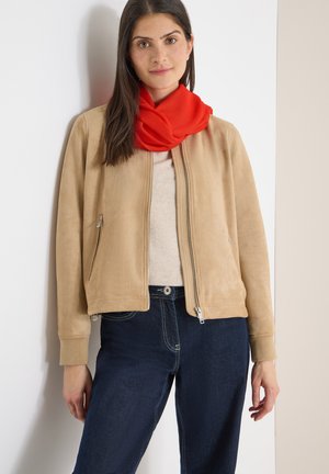 Joven mujer con cabello largo y oscuro, vestida con chaqueta de ante beige, bufanda roja, suéter claro y jeans azul oscuro, de pie contra una pared blanca.