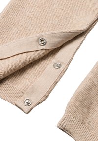 Beige gebreide stof met een gestructureerd patroon. Heeft drie metalen drukkers langs de opening en ribbelboorden aan de randen.