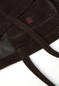 Sac fourre-tout en daim marron avec des accents en cuir, doté d'une poche zippée et de poignées cousues. Intérieur doublé, offrant une texture douce et un design durable.