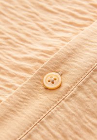 Tessuto beige con una texture arricciata, caratterizzato da un bottone giallo rotondo con quattro fori, cucito saldamente sul bordo.