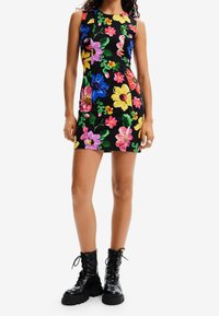 Robe sans manches à fleurs avec des fleurs multicolores vives sur un fond noir ; coupe ajustée avec un col rond. Associée à des bottes de combat noires.