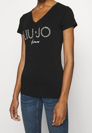 Persoon draagt een zwart V-hals T-shirt met de tekst "LIU·JO" en "dream" van strass-steentjes, gecombineerd met een blauwe jeans tegen een witte achtergrond.