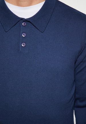 Cămașă polo bleumarin dintr-un material tricotat texturat, având un guler ascuțit și trei nasturi purpurii. Strat de bază este un tricou alb.