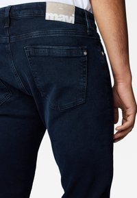 Close-up of a person wearing dark blue denim jeans with a visible back pocket and beige label reading "mavi."
