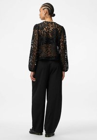 Svart blomstermønstrete blondetopp med lange ermer og åpen rygg. Kombinert med løstsittende svarte bukser og kraftige svarte sko.