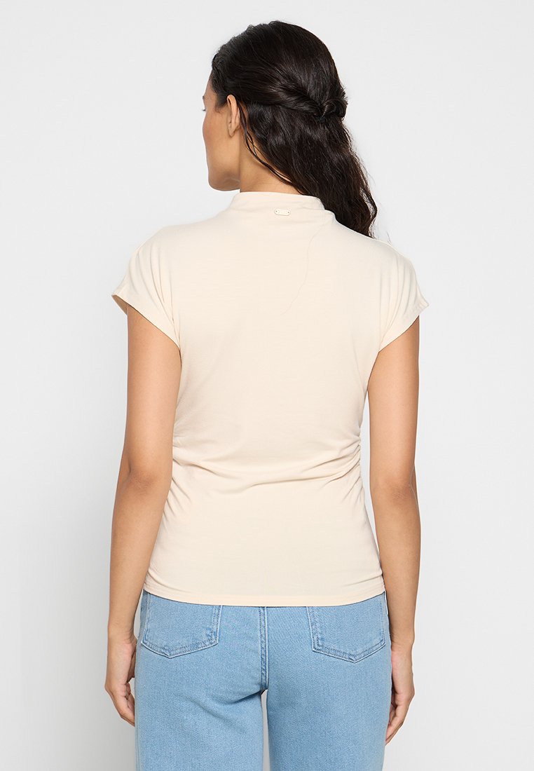 Beige Oberteil mit kurzen Ärmeln, tailliertem Schnitt, weicher Textur und hohem Kragen. Bemerkenswertes Detail: kleines Logo auf der Rückseite. Dazu eine Jeans in Blau.