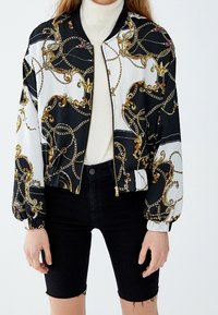 Veste bomber avec un motif noir et blanc orné de chaînes dorées. Fabriquée en matériau lisse, elle possède une fermeture éclair à l'avant et des poignets élastiques.
