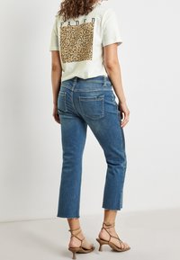 Žena v zkrácených modrých džínech s roztrhanými lemy, v béžových sandálech na podpatku s pásky a bílém tričku s květinovým motivem a textem "FADED" na zádech.