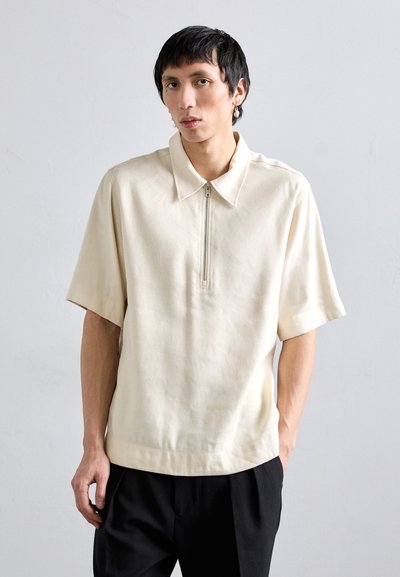 Beige Poloshirt mit kurzen Ärmeln, halbem Reißverschluss und Kragen, ausgestattet mit einer glatten Textur und lockerem Schnitt.