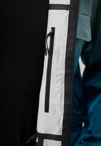 Veste blanche et noire avec une poche zippée sur la manche gauche, présentant un design épuré et des textures contrastantes. Aucune embellissement visible.