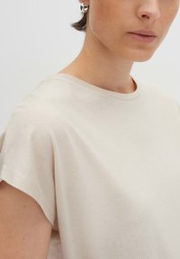 T-shirt in cotone beige con maniche corte, scollo rotondo e texture morbida, che presenta un design semplice e pulito.