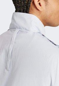 Leichte, weiße Jacke mit hohem Kragen, strukturiertem Stoff und einem schmalen Aufhängeriemen. Verfügt über kontrastierende Seitenelemente und Nähte.