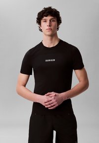 Ung mann med krøllete hår iført en svart Calvin Klein T-skjorte, står med hendene lett sammenfoldet mot en ensfarget grå bakgrunn.