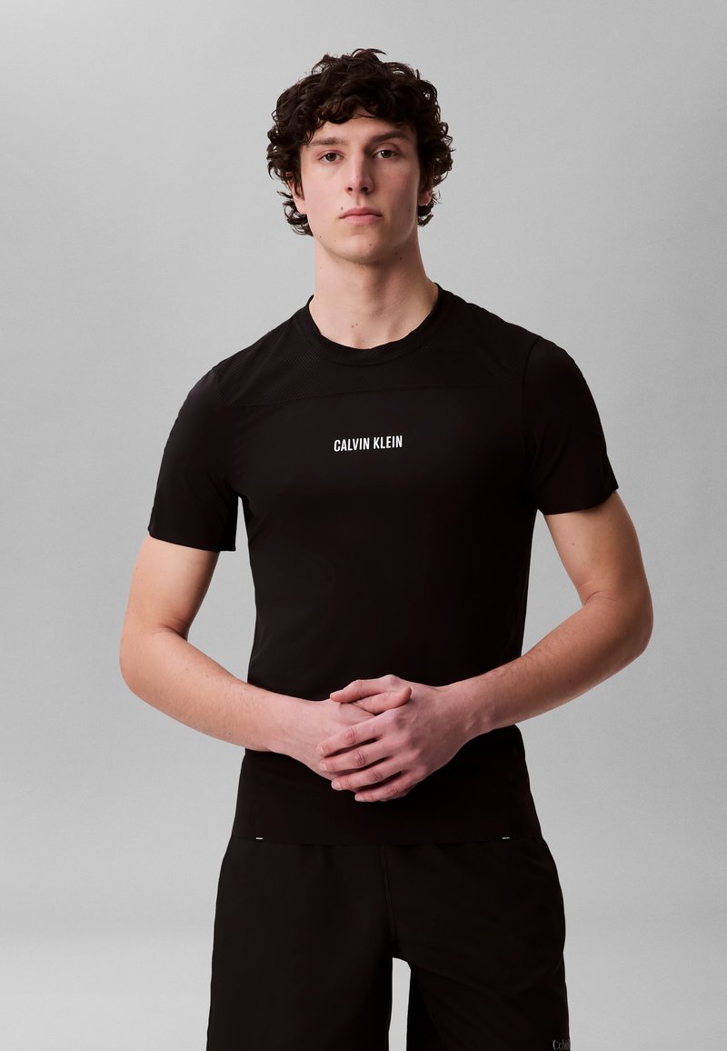 Ung mann med krøllete hår iført en svart Calvin Klein T-skjorte, står med hendene lett sammenfoldet mot en ensfarget grå bakgrunn.