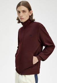 Giacca a zip bordeaux realizzata in tessuto liscio, con colletto alto, tasche laterali e un piccolo emblema sul lato sinistro del petto.