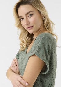 Green textured blouse with a v-neck, short sleeves, and openwork floral pattern. Model poses with arms crossed, showcasing sleeve design.