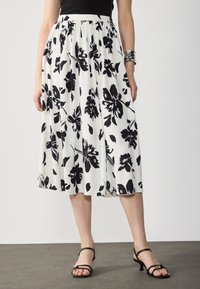 Jupe midi plissée blanche avec un motif floral noir. Dotée d'une taille élastique, d'un tissu léger et d'un design fluide. Sandales noires à lanières.