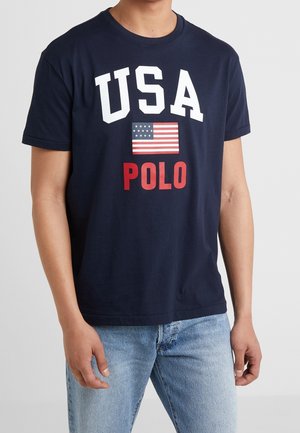 Personne portant un T-shirt bleu marine avec "USA", un drapeau américain et "POLO" imprimés en blanc et rouge, associé à un jean bleu clair.