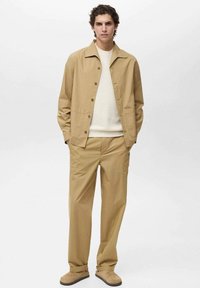 Beige leichte Jacke mit Druckknöpfen und zwei Fronttaschen, kombiniert mit lockeren beigen Hosen und einem cremefarbenen Strickpullover. Beigetöne Schuhe.