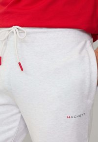 Sive pamukaste trenirke s bijelim žicom za podešavanje, crvenim vrhovima i bočnim džepovima. Sadrži "HACKETT" logo u crvenoj i sivoj boji na donjem dijelu nogavice.