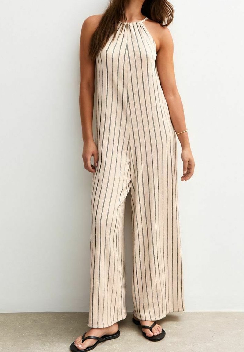 Femme portant une combinaison beige sans manches à jambes larges avec des rayures noires verticales et des tongs noires, debout contre un mur blanc uni.