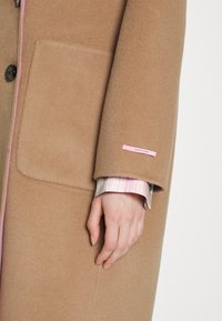 Bruine wollen jas met een zachte textuur, voorzien van een grote zak, roze accenten langs de randen en een label op de mouw. Gestreept shirt eronder.