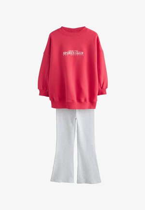 Raudonas per didelis džemperis su užrašu „SPORTS CLUB“, derinamas su šviesiai pilkomis platėjusiomis kelnėmis, pateiktas ant balto fono.