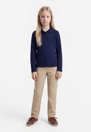 Szőke lány áll előre nézve, tengerészkék hosszú ujjú galléros pólót, bézs nadrágot és barna fűzős cipőt viselve fehér háttér előtt.