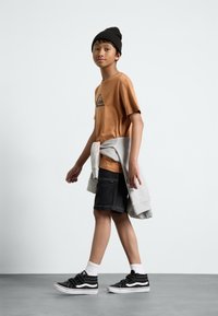 Camiseta de manga corta marrón con logo triangular negro, pantalones cortos negros, sudadera gris atada a la cintura, zapatillas negras con acentos blancos y calcetines blancos.