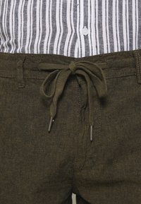 Pantaloni verde oliva con cordoncino e finitura testurizzata, abbinati a una camicia a righe verticali bianca e grigia chiara.