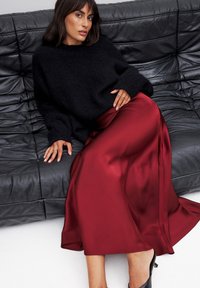 Schwarzer, flauschiger Pullover, roter Satinrock mit glatter Textur, getragen mit schwarzen High Heels, sitzt auf einer schwarzen Ledercouch.