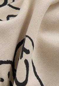Beżowa tkanina o miękkiej fakturze z wyrazistymi czarnymi abstrakcyjnymi wzorami. Materiał wydaje się gładki, z widocznymi szczegółami dzianiny.