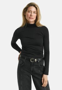 Blonde Frau in einem schwarzen Rollkragenoberteil und dunklen Jeans mit einem schwarzen Gürtel, die mit einer Hand auf der Hüfte vor einem weißen Hintergrund steht.