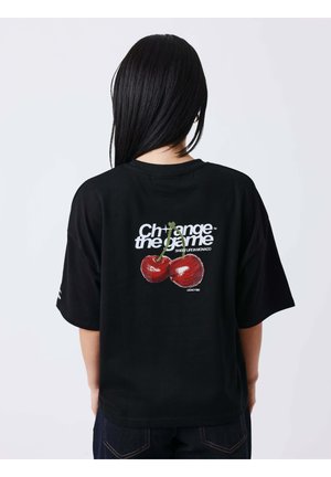 Persona con cabello largo y negro que lleva una camiseta negra con dos cerezas rojas y texto blanco "Change the game Sweet Life Monaco" en la parte trasera.