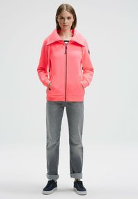 Pink zip-up hoodie with a high collar, featuring two front pockets and black accents on the shoulders, paired with gray rolled cuff jeans and navy shoes.