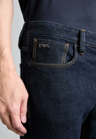 Jeans de mezclilla oscura con costuras naranjas, una etiqueta de metal en el bolsillo trasero y un diseño de pierna recta. Textura suave, sin patrones visibles.