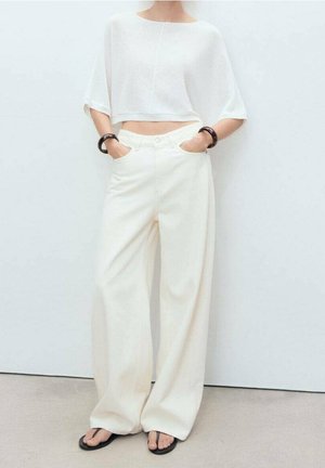 Person iført løstsiddende hvide bukser, en kort hvid top, sorte armbånd og sorte klipklappere, stående med hænderne i lommerne mod en ensfarvet væg.