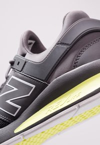 Černá a šedá sportovní obuv s polstrovaným límcem, texturovaným síťovaným svrškem, velkým reflexním logem a jasně žlutým detailem na podrážce.