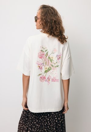 Kobieta z kręconymi włosami, w okularach przeciwsłonecznych, luźnej białej koszulce z różowym kwiatowym wzorem i napisem "BIZUU" na plecach oraz czarnej spódnicy w grochy.