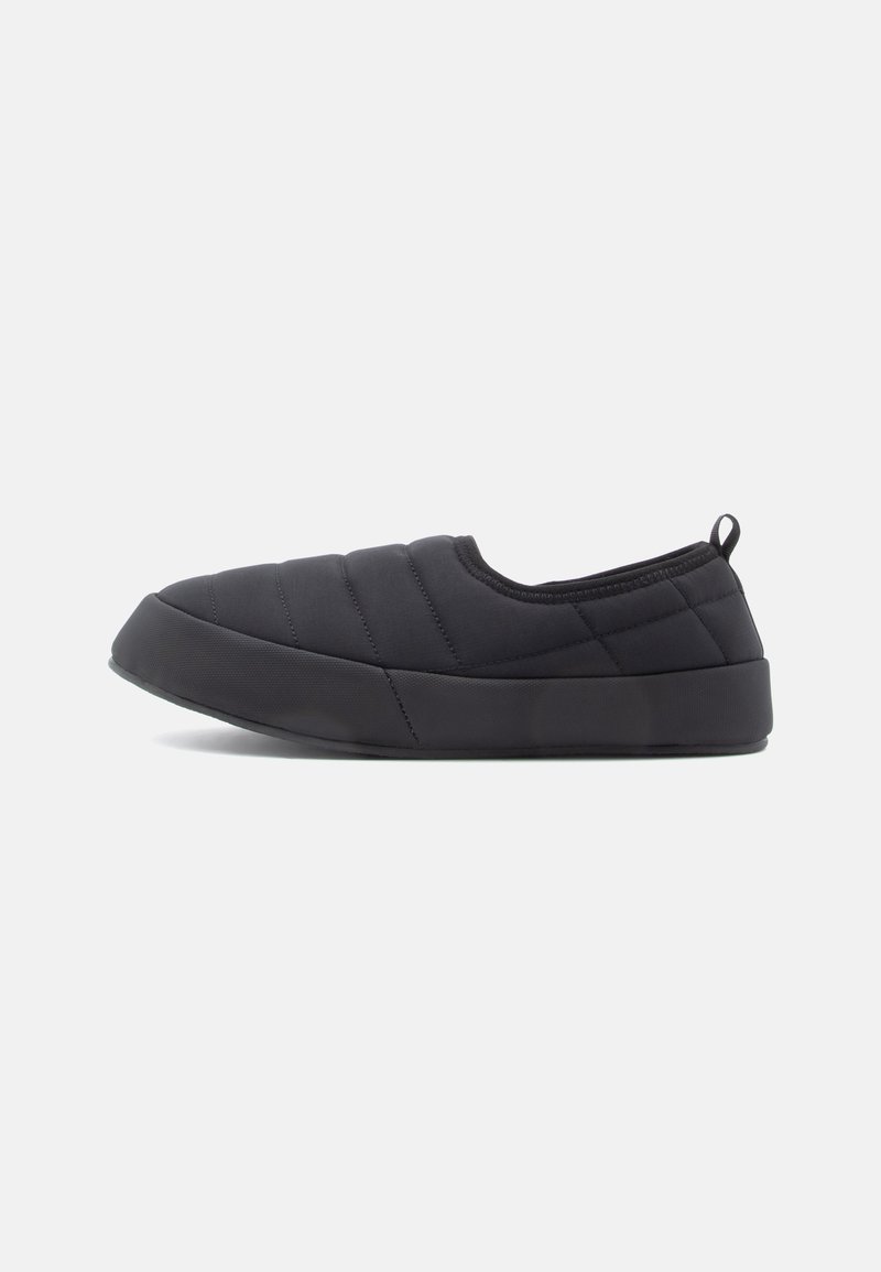 Chaussure noire matelassée à enfiler avec une semelle épaisse en caoutchouc et une languette au talon, présentée de profil sur un fond clair uni.