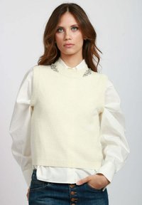 Gilet lavorato a maglia color crema con design senza maniche, caratterizzato da un colletto a coste e dei dettagli decorativi in argento sulle spalle. Camicia bianca sotto.